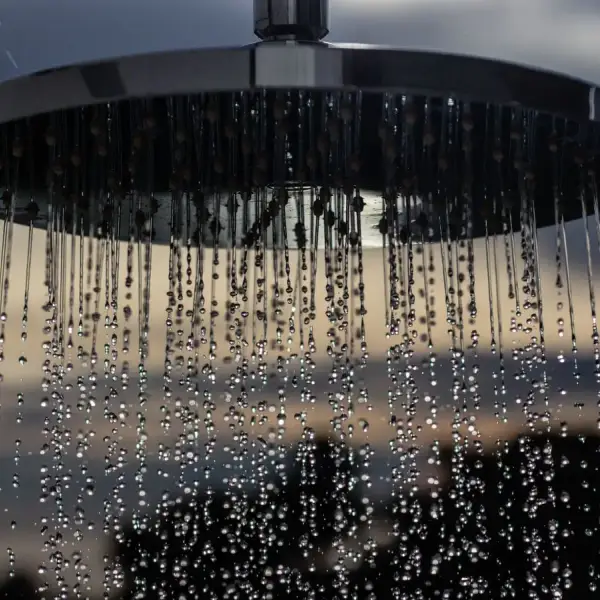 A detailed view of a shower head outdoors with drops flowing and a blurred sunset background.