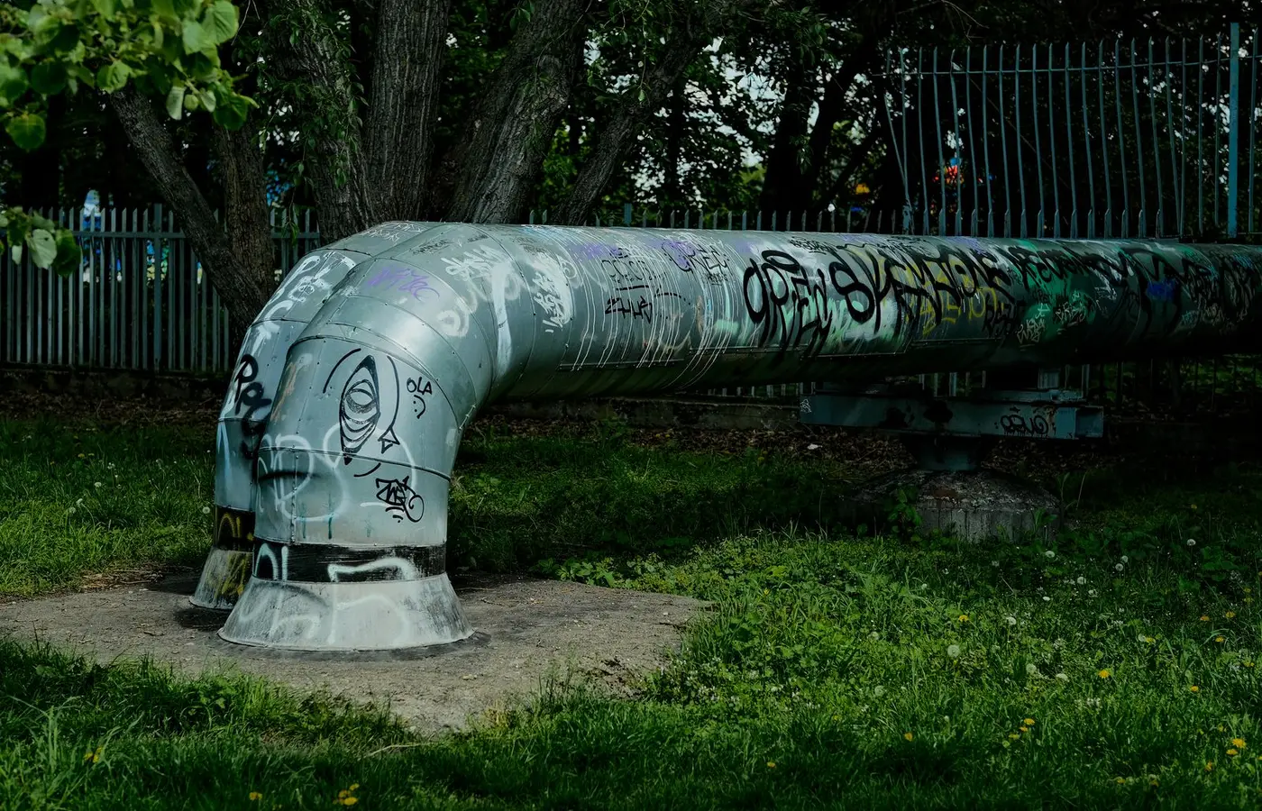 Large metallic pipe covered in graffiti amidst lush green grass and trees in a park setting.