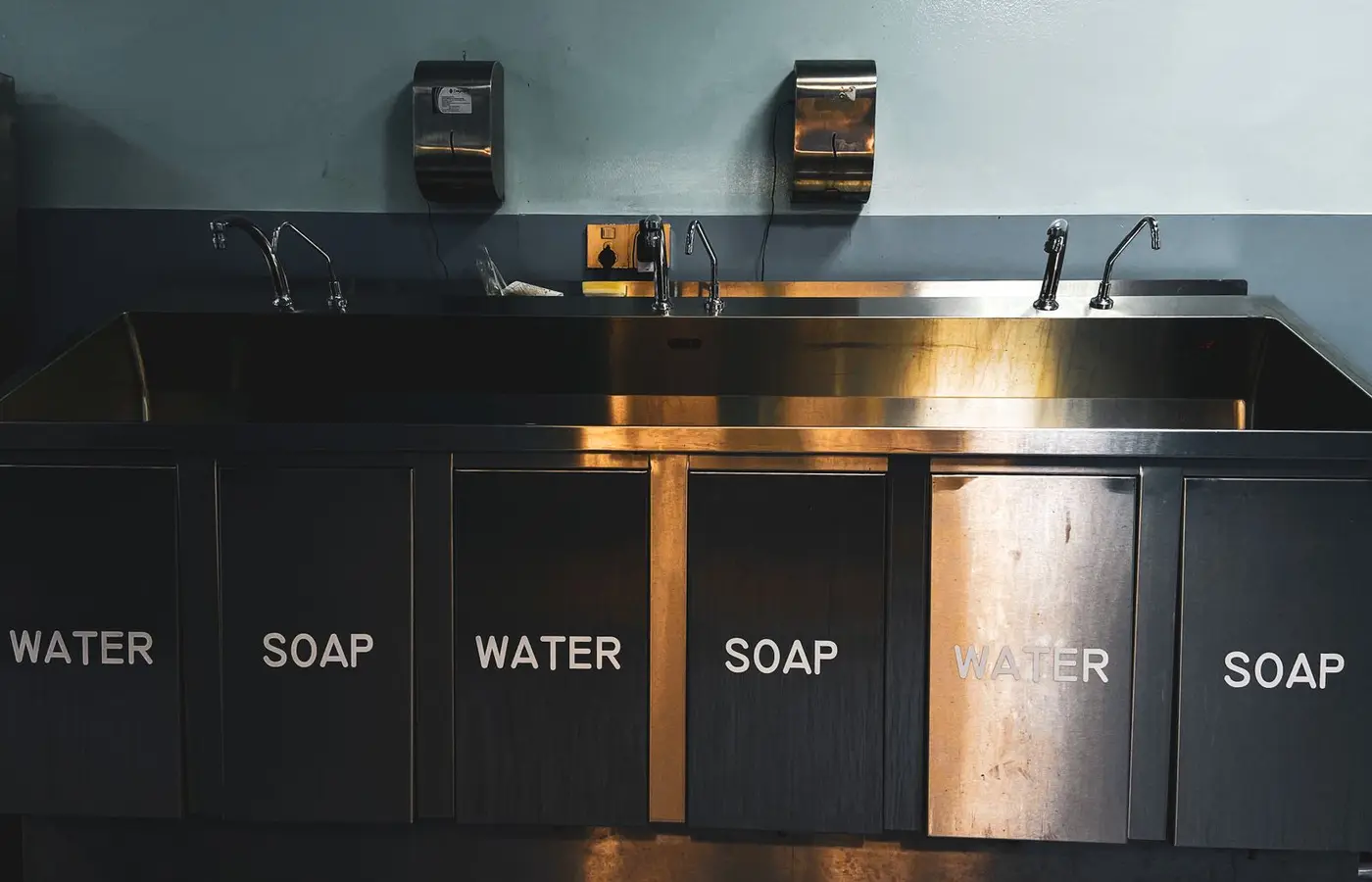 A stainless steel industrial kitchen sink with labeled compartments for water and soap.