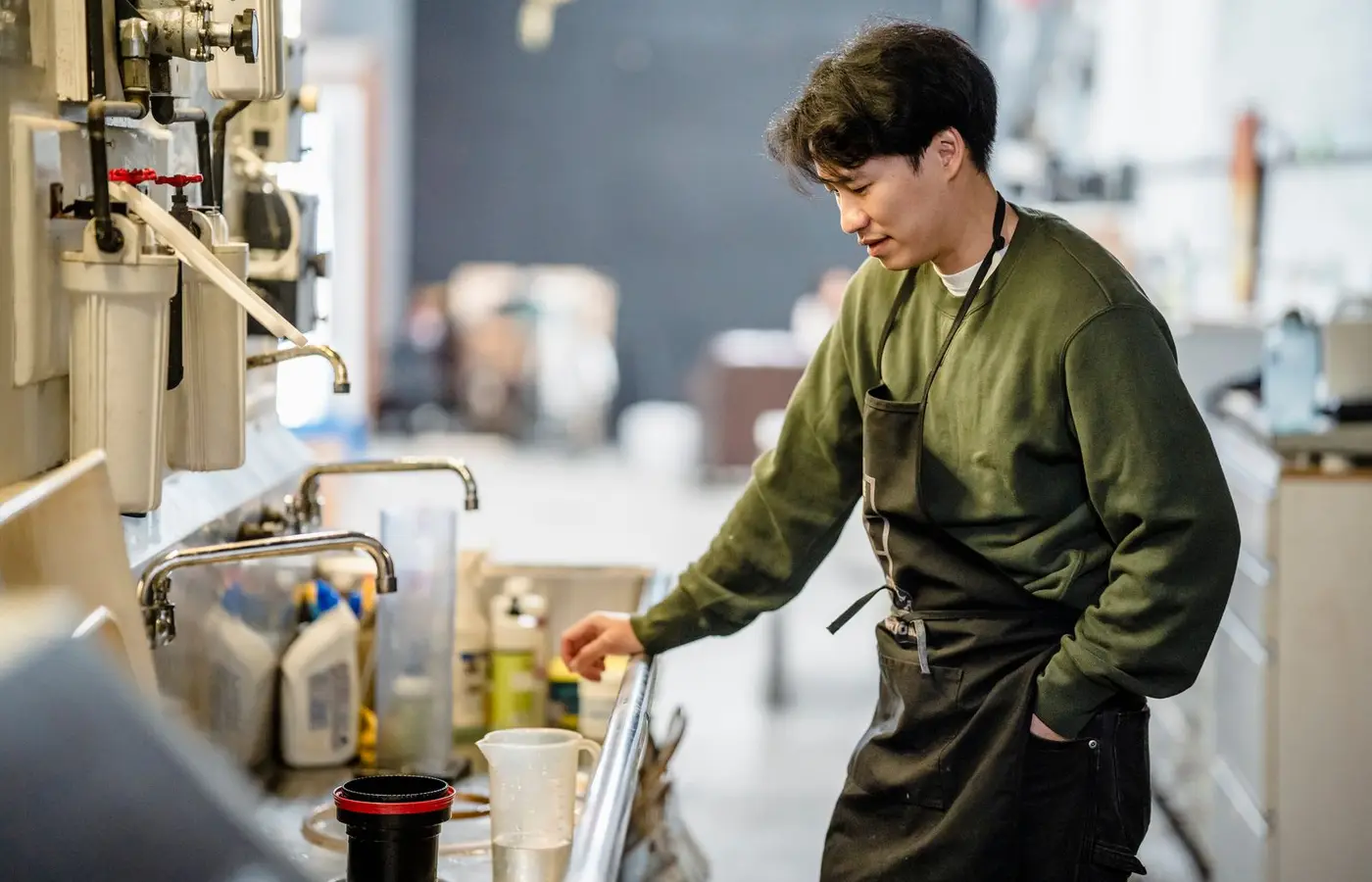 An Asian man in a commercial kitchen, wearing an apron and sweatshirt, standing by a sink.
