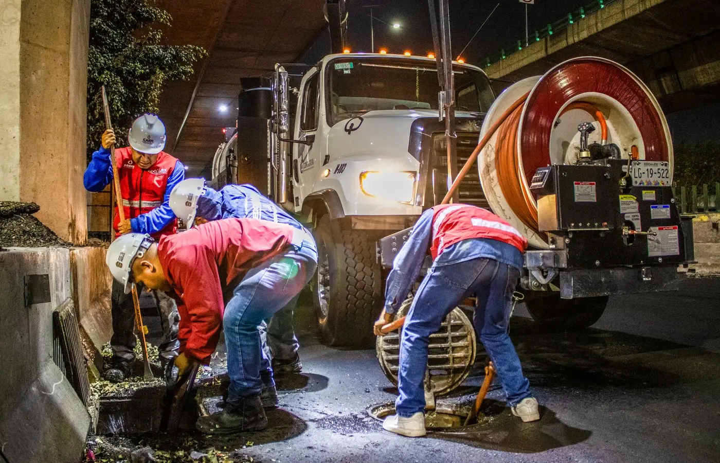 Street workers cleaning drainage at night with machinery under a bridge.