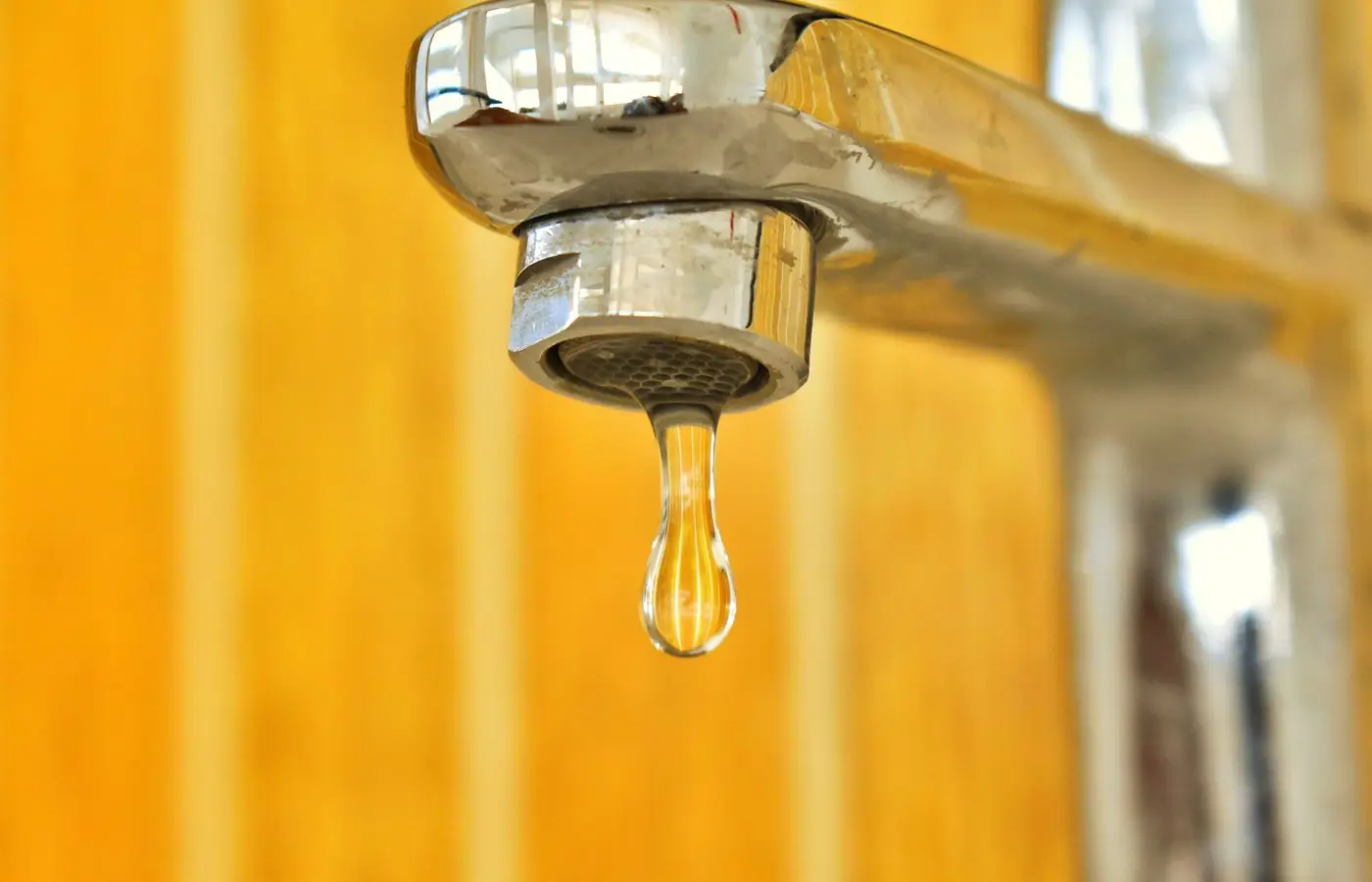 A close-up shot of a water droplet hanging from a stainless steel faucet, highlighting surface tension.